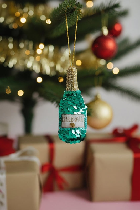 Green Sequin Bottle Ornament — A sequined green bottle-shaped Christmas ornament with a gold cap and hanging string, displayed in front of a blurred Christmas tree and wrapped gift boxes. — Zohricanada