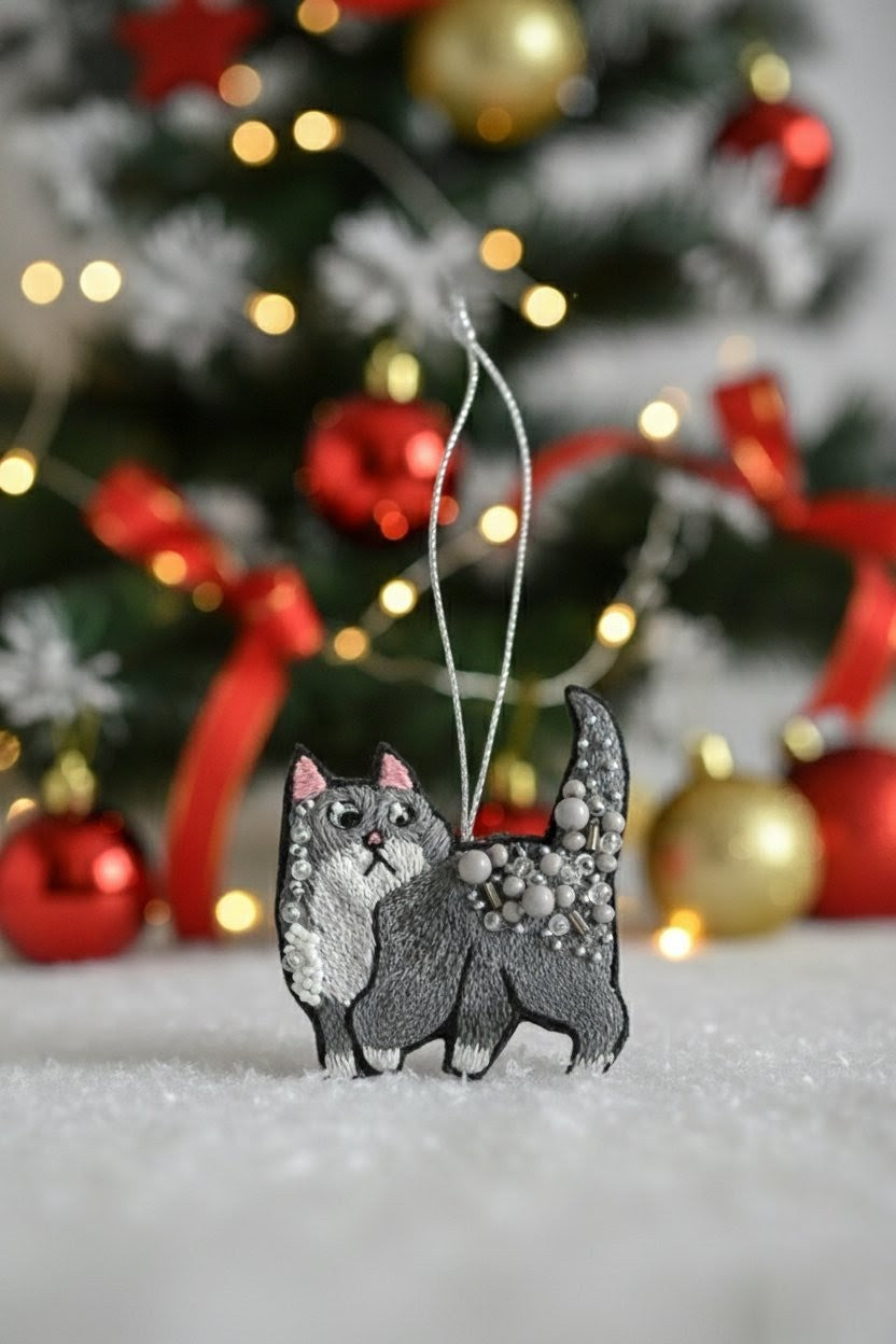 A gray and white cat-shaped Christmas ornament with glittery details hangs from a silver string in front of a blurred decorated Christmas tree with red and gold ornaments.