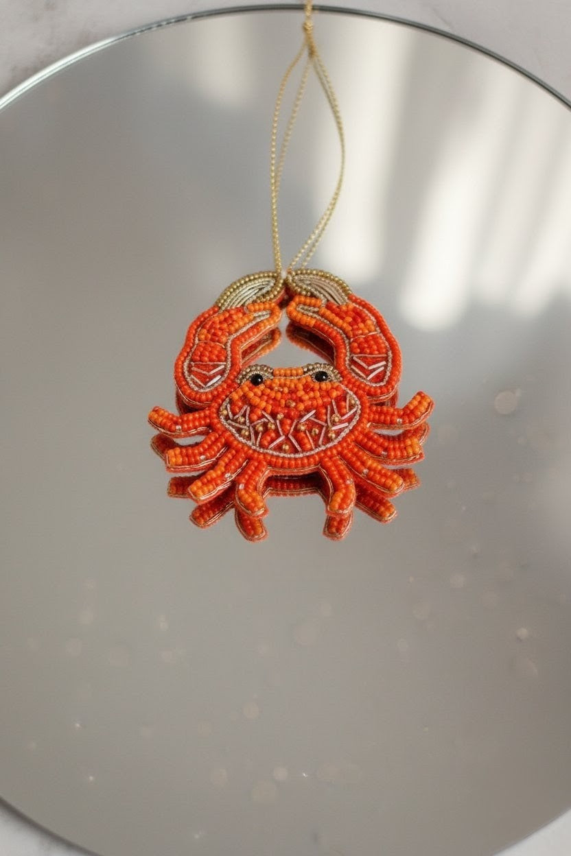A small orange beaded crab ornament with a gold hanging string, photographed against a reflective silver surface.