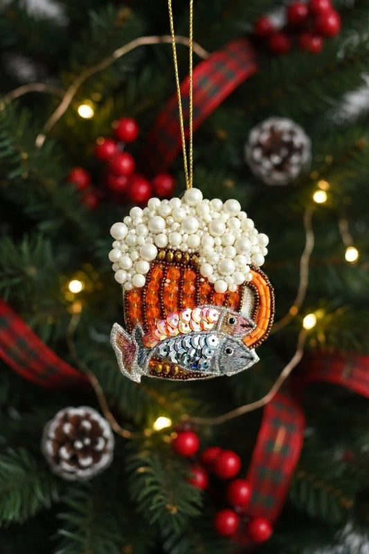 Fish Beer Mug Ornament — A Christmas ornament shaped like a mug filled with white beer foam and decorated with orange and silver beads, hanging on a decorated Christmas tree with red berries, pinecones, and warm string lights. — Zohricanada