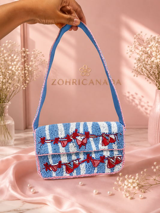 Seaside Bikini Handmade Beaded Shoulder Bag — A hand holds a small blue and white striped beaded handbag with red bow embellishments and a blue shoulder strap, set against a pink backdrop with the brand name ZOHRICANADA visible. — Zohricanada